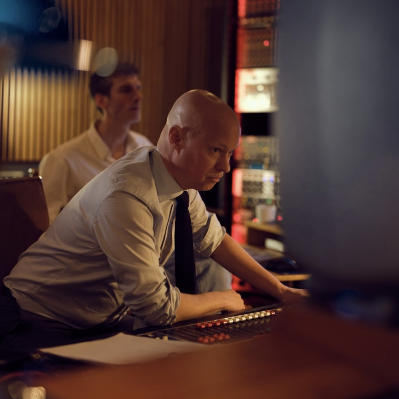 An engineer at the mixing desk mid-session, a colleague in soft focus behind, a rack of outboard gear glowing warm red in the background